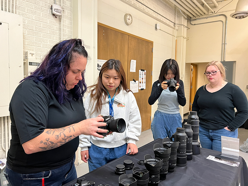 A woman with dark hair in a dark shirt holding and looking at a camera, showing it to a female student while standing behind a table filled with other lenses, and two other students standing next to the table looking at camera equipment