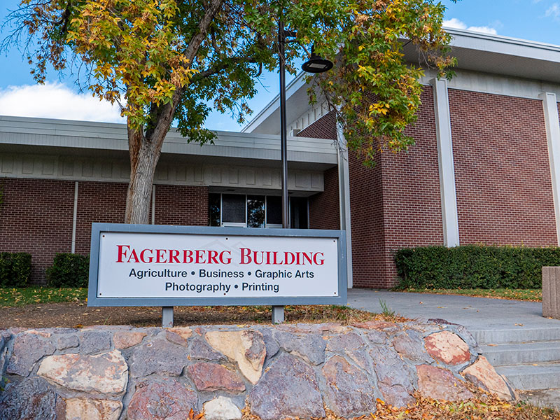 Outside the Fagerberg Building with a sign with red lettering on top of a stone shelf with red-bricked building, fall colors, and a blue sky in the background