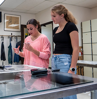 Two female high school students examine a printing project standing over a table covered in printmaking materials