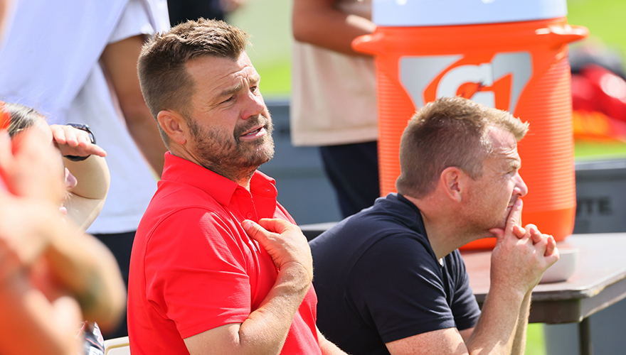 Two male soccer coaches sitting on a bench on the sideline, one with a beard in a red shirt with his hand on his chest, the other in a gray shirt leaning forward