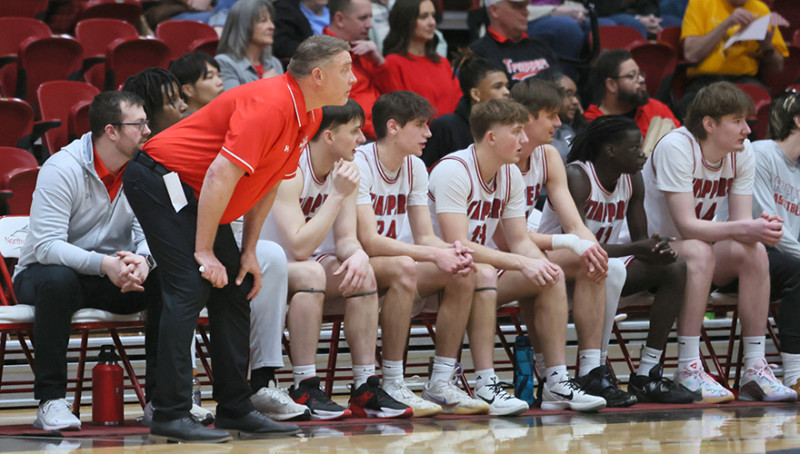 A men's basketball coach in red shirt with his hands on his knees looking across the court with several players sitting in chairs behind him