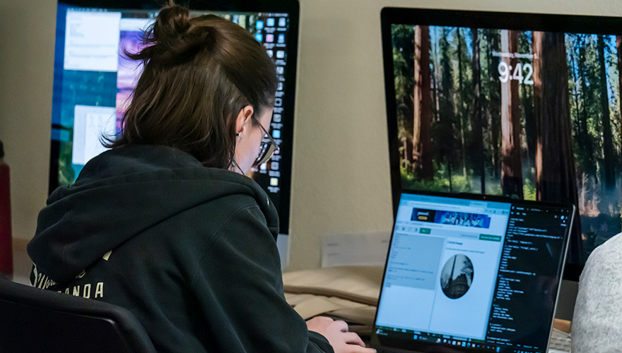 Female student typing on a laptop with graphics on a screen in front of her and larger monitors in the background
