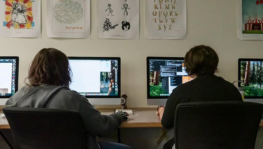 Two students sitting facing computer screens in a classroom with images on the wall above them