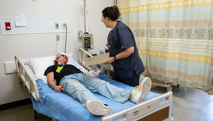 A female nursing student holding a manila folder checking vital signs on a monitor of a patient in bed