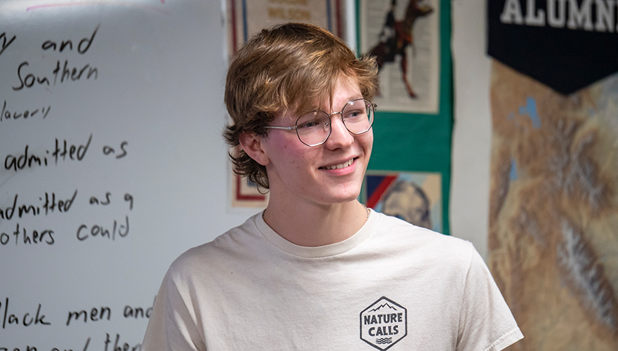 Male college student wearing glasses, smiling and standing in front of a classroom and white board