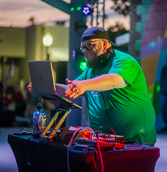 A male DJ wearing a bright green shirt playing music on a laptop outdoors at sunset