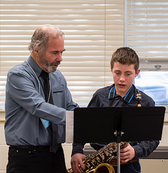 A male instructor on the left and young student on the right standing behind a music stand holding a saxophone
