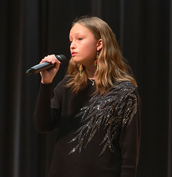 A close-up of a young female student on stage singing into a microphone she's holding in front of black drapes