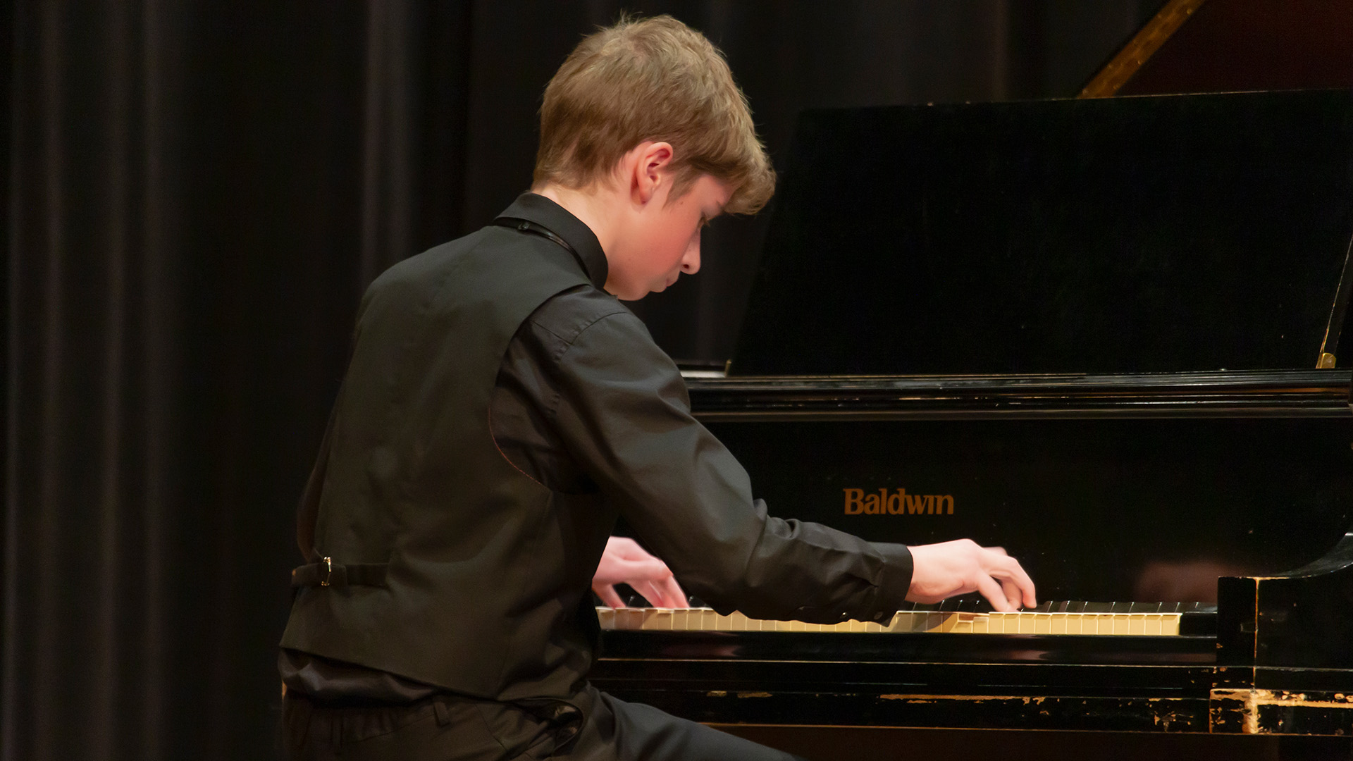 A young male student in black playing the piano on stage
