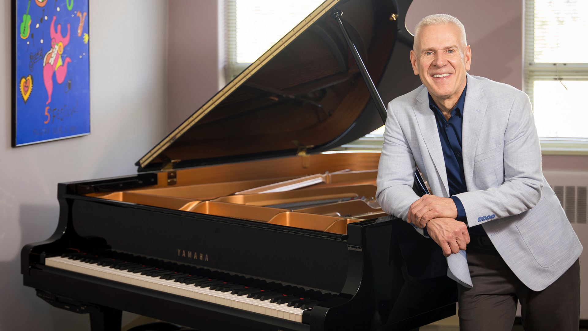 A man with short gray hair leaning on a grand piano in a studio setting with artwork hanging on the wall
