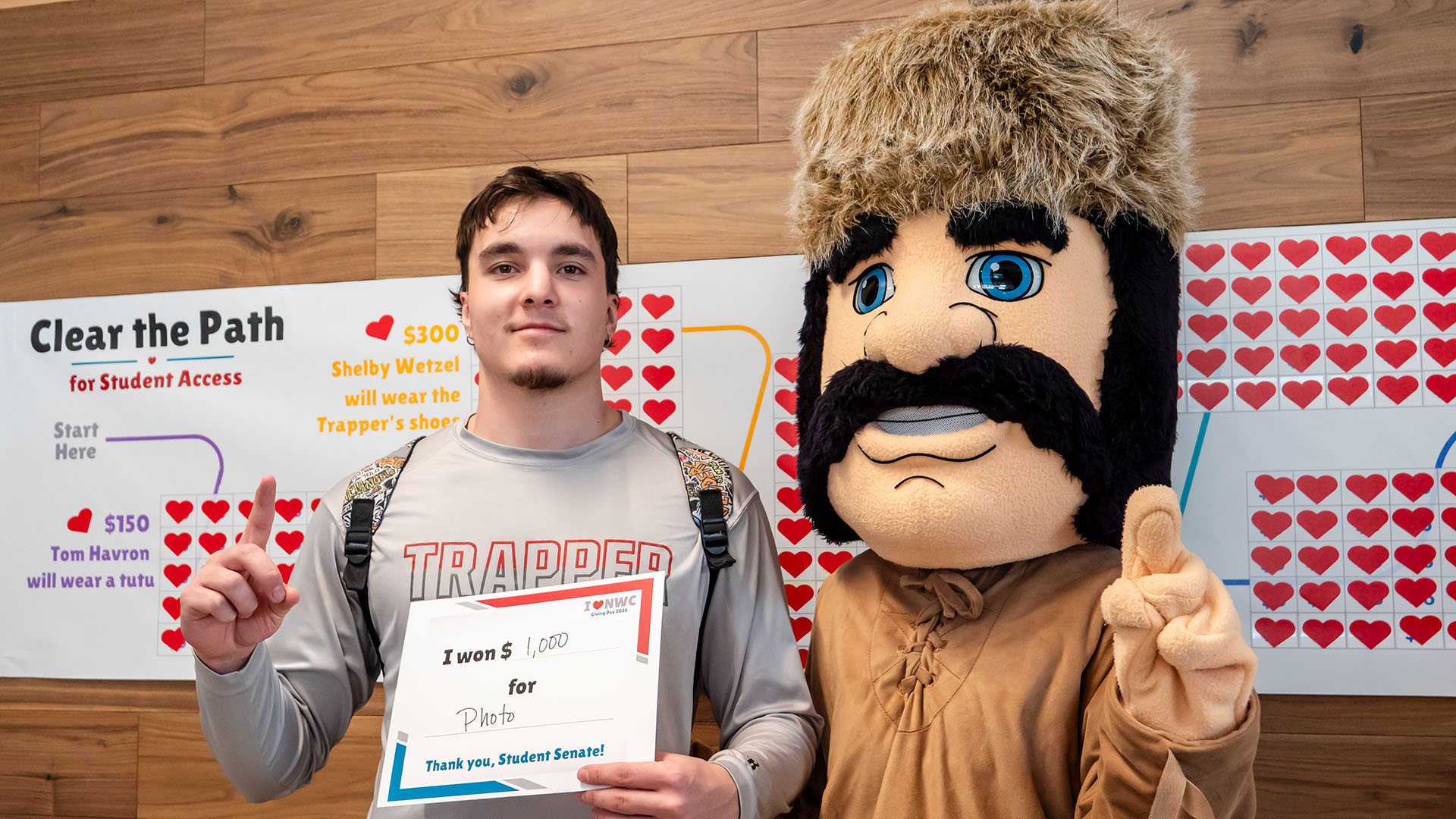 A male student holding a certificate standing next to the Trapper mascot in front of a banner filled with hearts