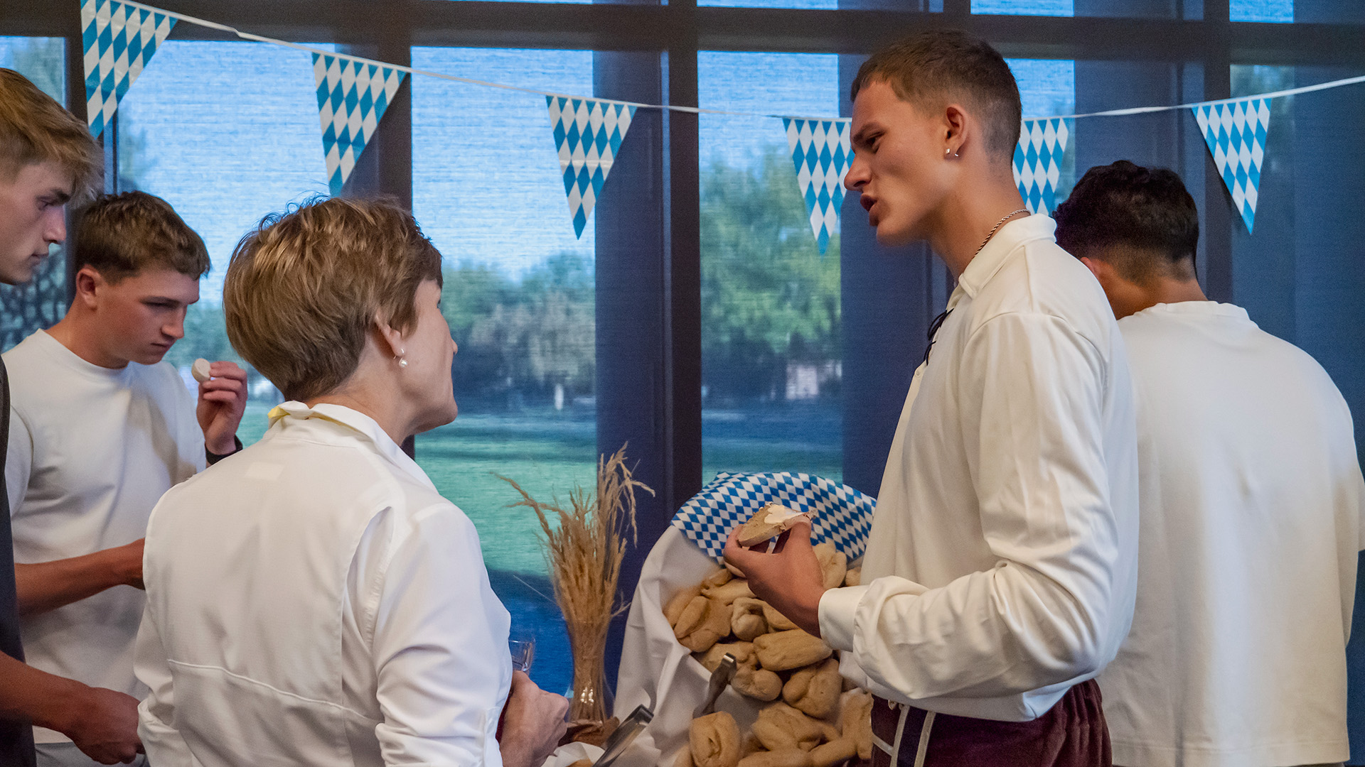 A woman on the left and man on the right dressed in traditional German clothing talking in front of a table full of bread with windows opening onto green grass and trees beyond