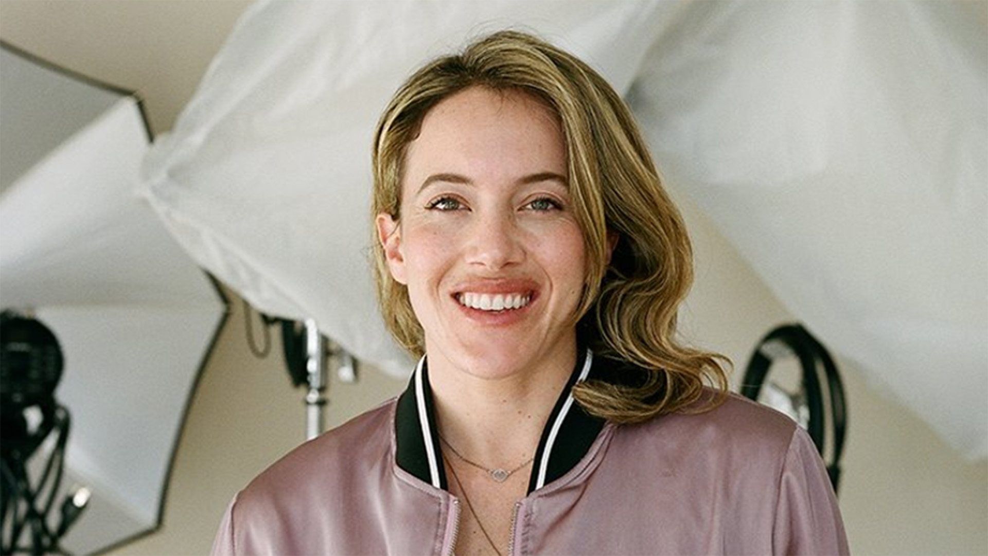 A woman with dark blond hair wearing a light purple shirt with a black collar standing in a photo studio