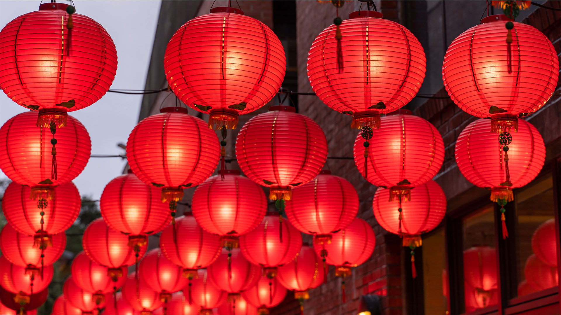 A large number of light-filled red orbs hanging above a street with a brick wall and windows in the background