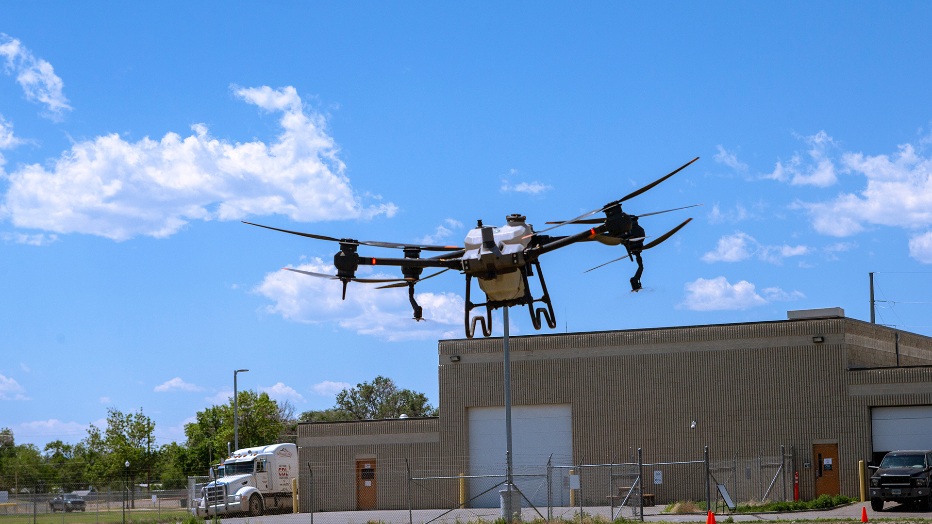 A drone hovering in the air against a partly cloudy blue sky with a tan colored building in the background blow