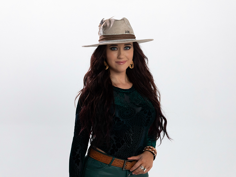 A female singer with long styled black hear wearing a long-sleeved black shirt, blue jeans, and a hat