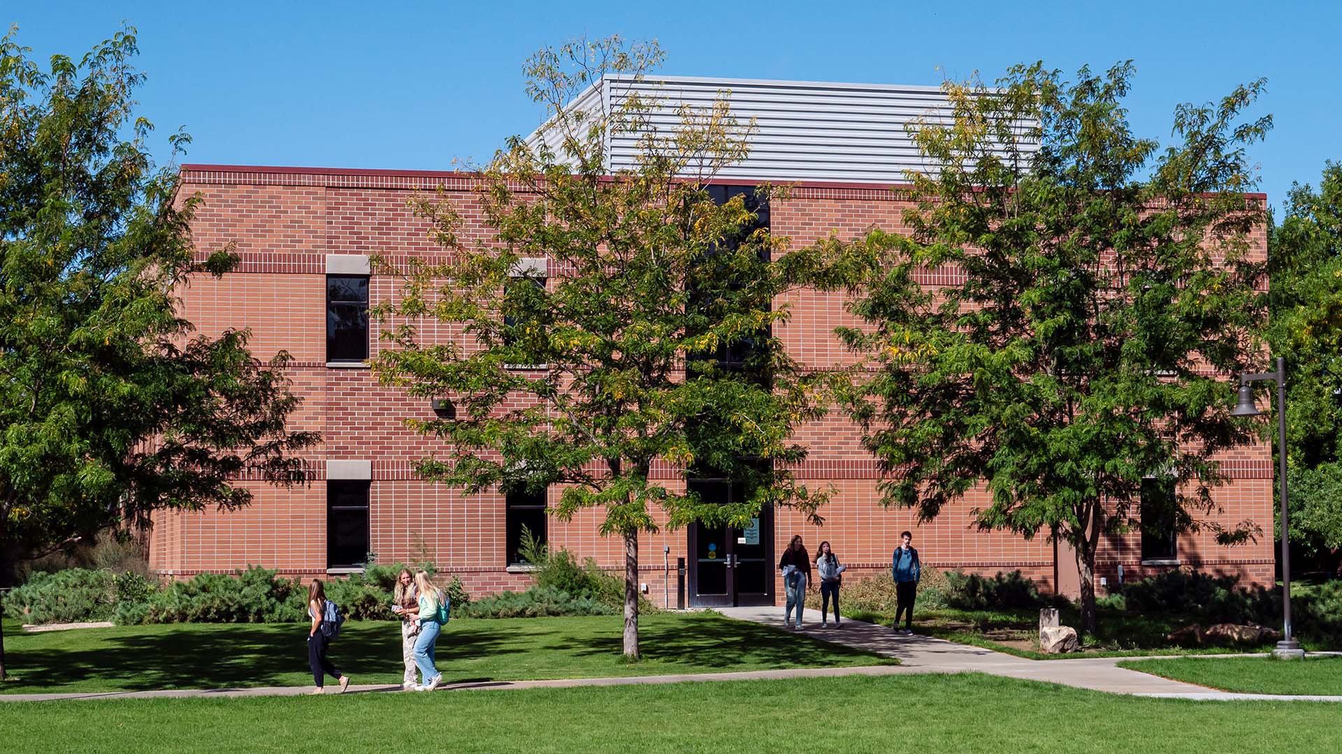 Students walking on the sidewalks between trees outside a two-story brick building with windows on a sunny September day