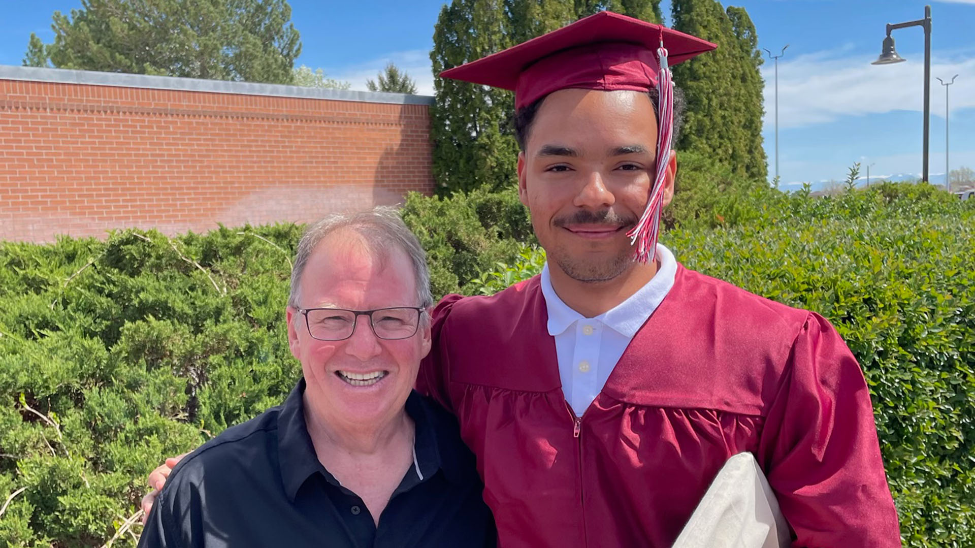 An older male with glasses wearing a black shirt standing next to a taller younger male in red cap and gown, both in front of hedges and a red brick building on a sunny day