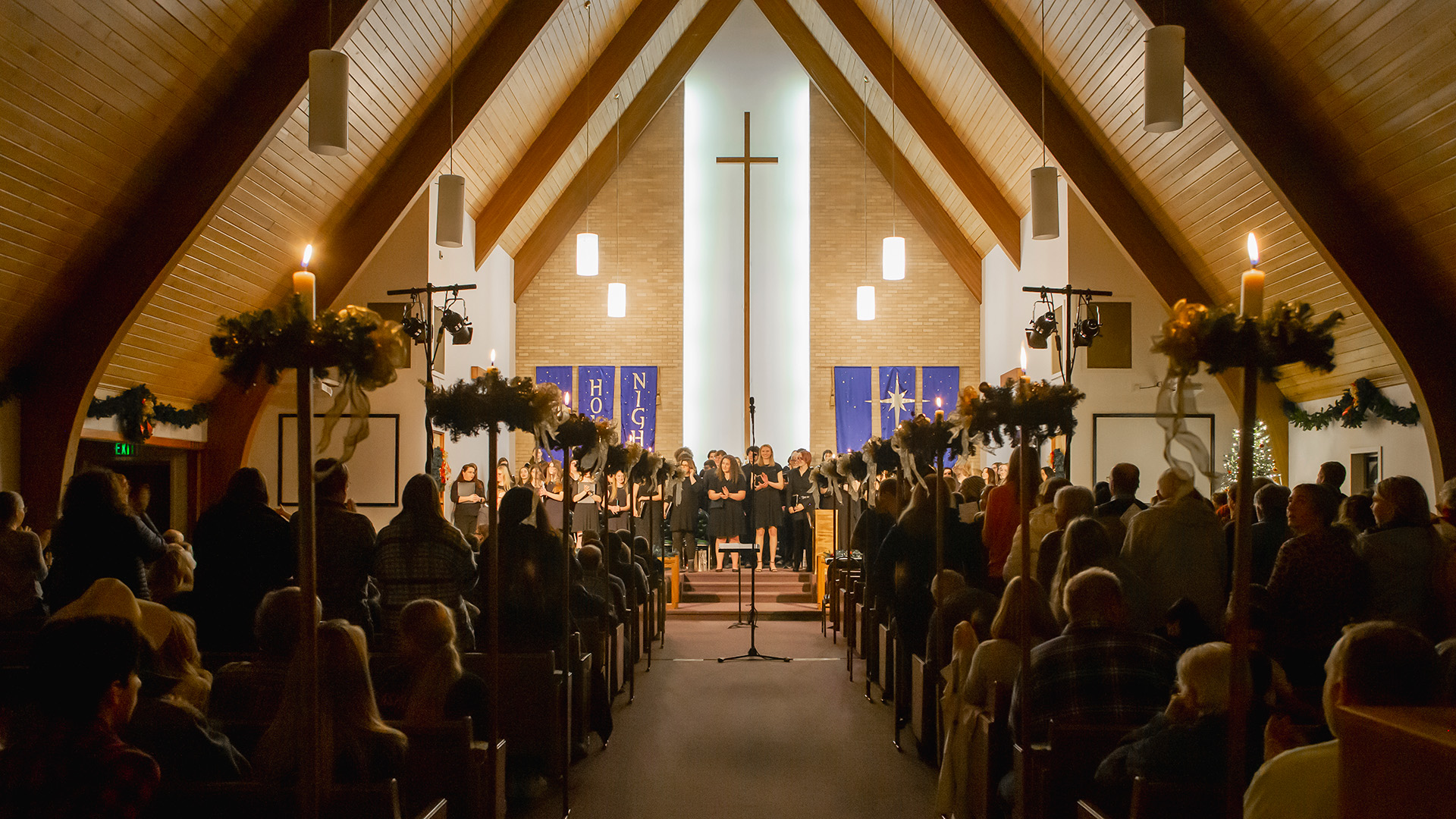 Looking down the center aisle of a dimly lit church at Christmas time with arched wooden beams overhead, pews filled with people, lit candles in stands on both sides of the aisle, with a choir singing beneath a large cross
