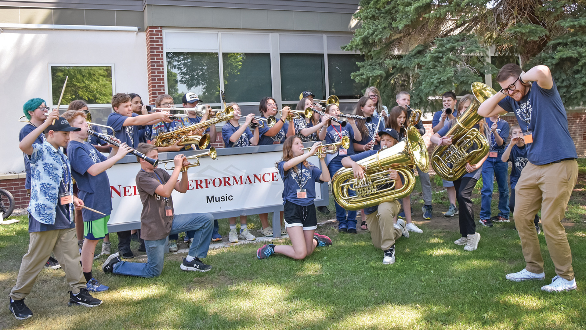 A group of young students outside a building playing instruments loudly toward a music teacher covering his ears