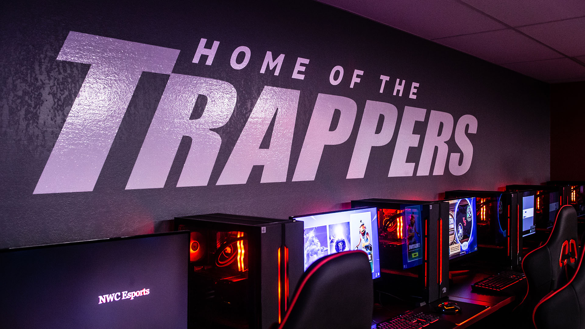 A dark wall with the words Home of the Trappers painted in white above computers, monitors, and gaming chairs